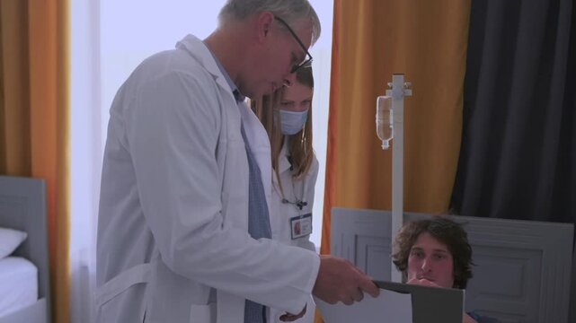 Group of doctors in white coats and masks gather around patient bed, making notes on tablets and papers while collecting medical anamnesis and discussing treatment options.