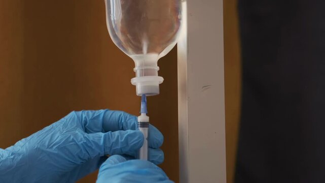 Gloved medical worker adjusting IV drip and filling syringe from infusion bottle. Close-up, nurse hands performing intravenous procedure in hospital environment. Professional healthcare treatment.