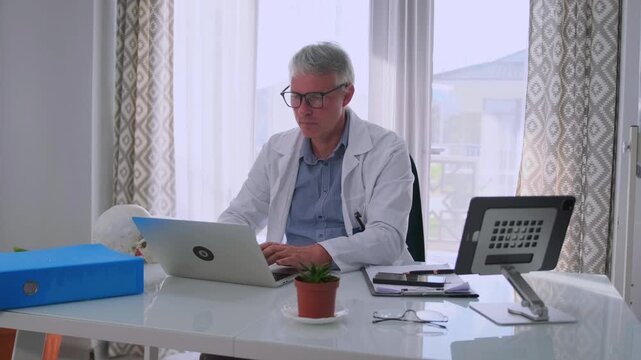 Male doctor in white coat and glasses working on laptop at modern medical office. Physician concentrated on typing and analyzing patient data, sitting at desk with documents and medical equipment.