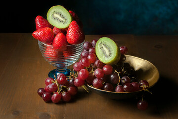 Still life photography with fruits, strawberries, grapes, and kiwis on an old wooden table with a dark blue background. Decorative photograph for framing.