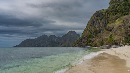 A beautiful tropical beach without people. The waves of the turquoise ocean foam and spread across the sandy beach. Boulders near the water. Bangka boat is moored far away. Mountains, blue sky, clouds