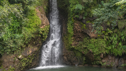 A picturesque tropical waterfall. Streams of white water flow down a sheer cliff into a turquoise lake. Thickets of green vegetation on the slope. Mauritius. La Vallée Des Couleurs Nature Park 