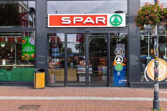 A bright, modern Spar convenience store in Arnhem, Netherlands, on a sunny day. The storefront features a large, red and green logo, glass doors, and windows displaying products.