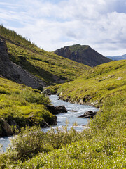 A winding river flows through a vibrant green valley under a partly cloudy sky. Lush hills rise on both sides, with distant mountains and a group of hikers visible on a path.