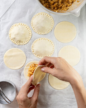Homemade apple and caramel mini pies preparation &mdash; dough cut-outs on bright marble with filling and hands sealing the top layer. Clean high key process shot with copy space.
