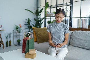 Asian woman sitting on sofa with shopping bags on table her using smartphone shopping online in living room at home