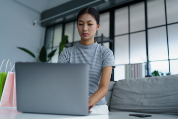Asian woman sitting on sofa with shopping bags on table her using laptop shopping online in living room at home