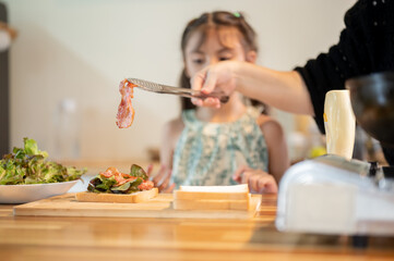 Asian daughter looking at mother putting bacon on bread making sandwich at kitchen cooking counter.
