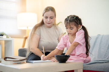 Caucasian mother looking at daughter holds pencil doing homework at table aside sofa in living room.