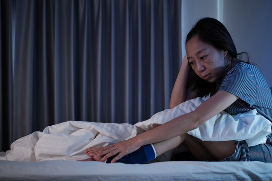 Female Asian middle-aged with ankle support is sleepless, has restless leg syndrome, is stressed, awake at night, with insomnia illness, mature and menopausal woman, and healthcare lifestyle problems.