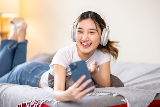 Fototapeta Happy asian woman wear headphones video calling talking on phone while lying on sofa bed in a room.