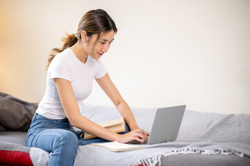 Naklejka premium Asian woman looking and typing on laptop aside books while sitting on sofa bed in a living room.