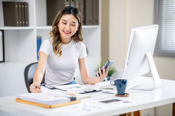 Asian woman office worker holding phone and looking at document while sitting at computer table.