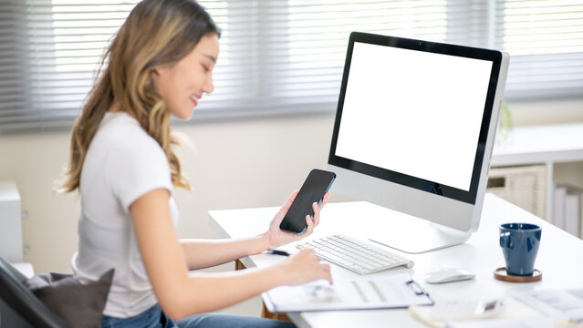 Asian woman office worker holding phone looking at clipboard and sitting with blank screen computer.