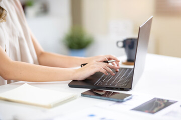 Fototapeta premium Close up of a woman office worker's hands holding pen and typing on laptop at white table in office.