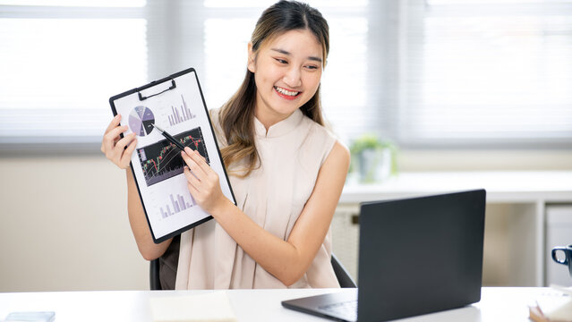 Pretty woman office worker holding a pen pointing on clipboard in front of laptop at table in office