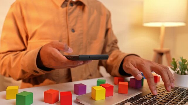 An office employee types on a phone while using a laptop surrounded by scattered colorful blocks, symbolizing inefficient multitasking, lack of focus, and the struggle to maintain productivity.
