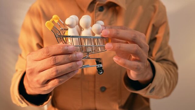 A person places wooden figurines into a small shopping cart, symbolizing sustainable human resource allocation, while also reflecting consumer behavior analysis and strategic workforce planning.

