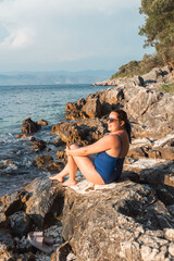 Woman in blue swimsuit sitting on coastal rocks, gazing at sea at sunset