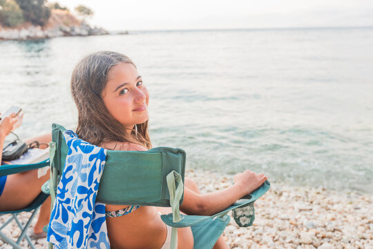 Teenage girl with long brown hair sits on a green beach chair by the rocky shore, smiling softly at the camera