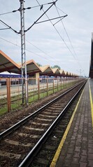 Empty railway tracks at a train station with patterned roofs and electric lines under a cloudy sky