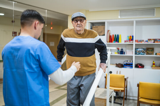 Senior man receiving physical therapy, walking with parallel bars, supported by a caregiver in a rehabilitation center