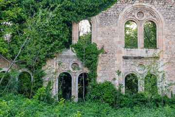 Overgrown facade of ancient abandoned stone building with arched windows and creeping ivy. Haunted ruined house covered with green plants. Post apocalyptic, creepy and spooky, romantic ruin concept