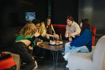 A lively scene of diverse friends sitting at a table indoors, sharing food and drinks, showcasing enjoyable social moments and friendship connections.