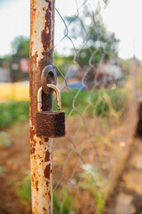 There is a lock on a metal post. The lock is rusted, against the background of a chain-link fence. Country fence.