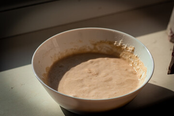 A white ceramic bowl containing a bubbly, active sourdough starter, placed on a windowsill and...