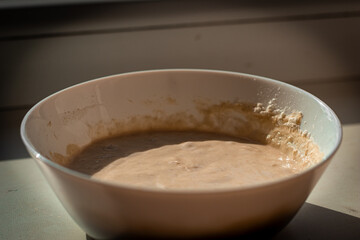 A white ceramic bowl containing a bubbly, active sourdough starter, placed on a windowsill and illuminated by natural sunlight. The mixture has a thick, frothy texture, indicating fermentation.