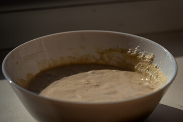 A white ceramic bowl containing a bubbly, active sourdough starter, placed on a windowsill and illuminated by natural sunlight. The mixture has a thick, frothy texture, indicating fermentation.