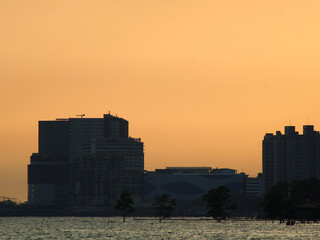 Modern skyscraper silhouettes on the water's edge at dusk, with a golden orange sky and calm water, creating a serene urban coastal view.