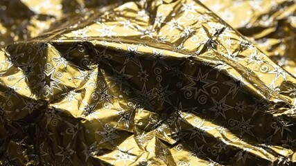 A close-up of crumpled gold foil with a reflective surface decorated with a repeating pattern of silver stars and spirals - Powered by Adobe
