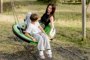 Mother and son sitting together on green swing at playground, enjoying tender family moment in nature on sunny day