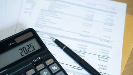 A black calculator showing 2025 placed on top of financial documents with a black pen nearby. The...