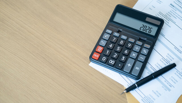A black calculator displaying 2026 with a fountain pen and financial documents on a wooden desk. The scene reflects careful accounting and strategic planning for financial security.