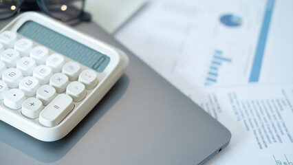 A white calculator with large buttons rests on a gray laptop beside financial documents with blue charts on a wooden desk symbolizing finance planning and analysis.