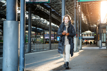 Naklejka premium Confident middle aged woman in winter coat walking on snowy train platform with phone, urban commuting, cold season lifestyle in bright daylight.
