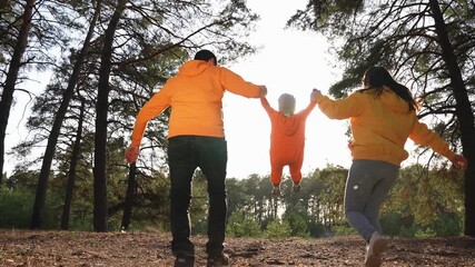 Parents swing child between hands in sunlit pine forest path with dry leaves on ground, family walk and laugh, parent child bond shows nature play and outdoor together, playful love and joy visible