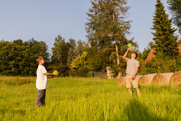 A father and son are spending quality time outdoors, playing catch with colorful balls. It is a warm, sunny day, and they are surrounded by lush greenery and hay bales