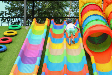 A young Asian boy in a white shirt and cap climbs a steep, multi-colored slide at a playground, showing determination and a sense of adventure.