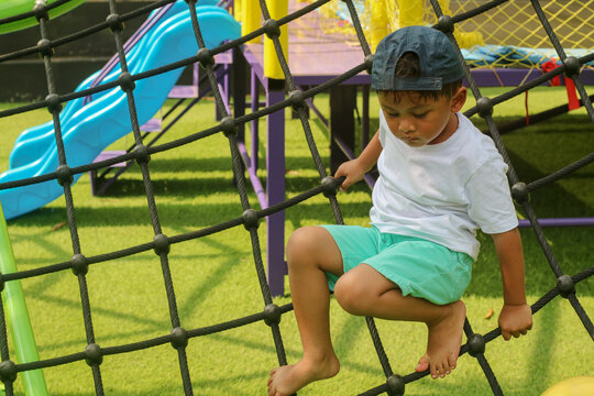 A young Asian boy with a cap sits on a black rope net at a playground, concentrating as he holds on. This image captures a moment of childhood determination and adventure.