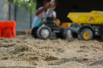 Bright toy dump truck rests in focus at an outdoor playground, with children playfully engaging with sand toys out of focus in the background during a sunny day.