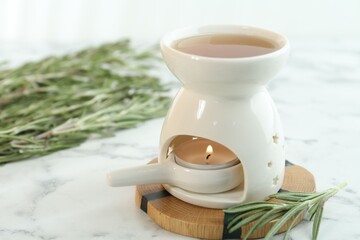 Aroma lamp with burning candle and rosemary on white marble table, closeup