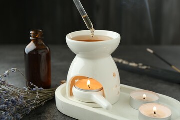 Dripping essential oil from pipette into aroma lamp with burning candle at grey textured table, closeup