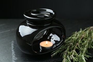 Aroma lamp with burning candle and rosemary on dark textured table against black background, closeup