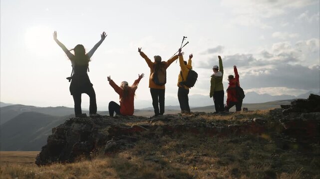 A group of diverse tourists trekkers stands together at the summit of a mountain, raising their arms in celebration of the end of their climb. The backdrop is a mountain range and sunset sky