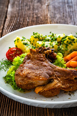 Baked veal cutlet with bone, boiled potatoes, mini carrots and green peas on wooden table