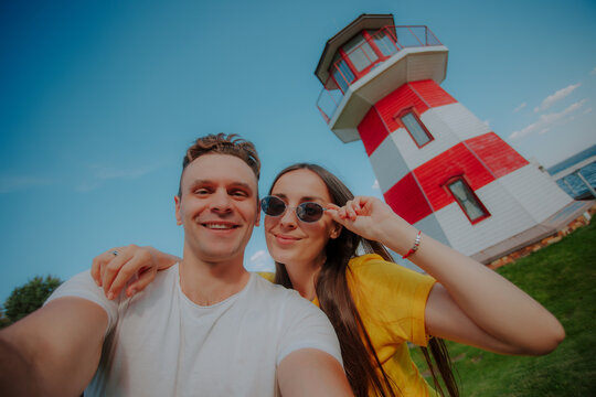 Happy couple taking a cheerful selfie in front of a red and white lighthouse on a sunny day, symbolizing love, travel, vacation, and joyful summer memories.
